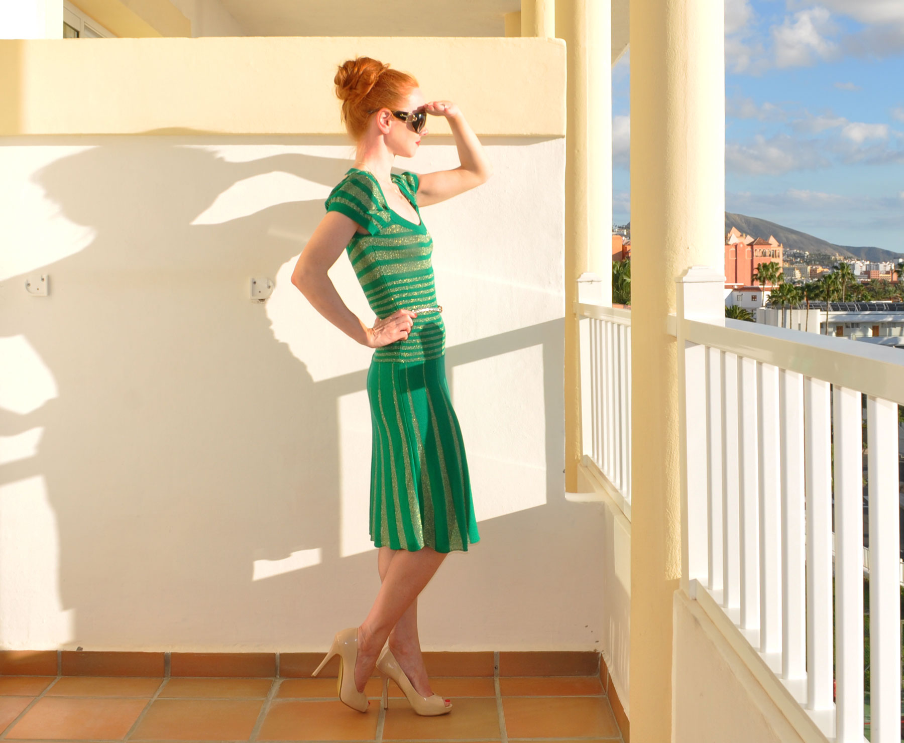 Amber wears a green and gold dress with high heeled shoes and shades her eyes from the sun as she looks out from her hotel balcony in Tenerife