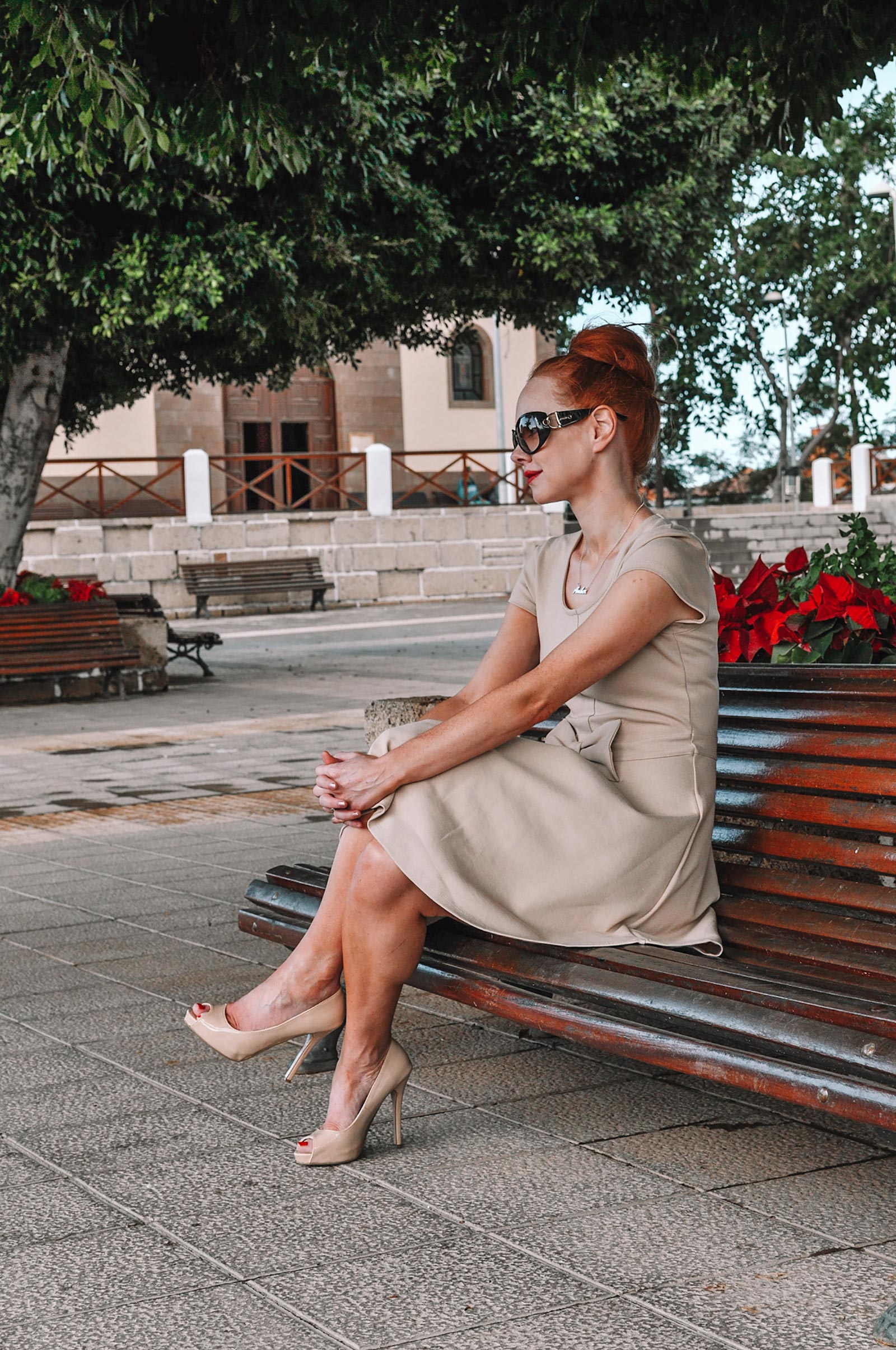 Amber sits on a park bench in a pretty Spanish square. She is wearing a beige mini dress, high heels, and dark sunglasses, with her hair up 