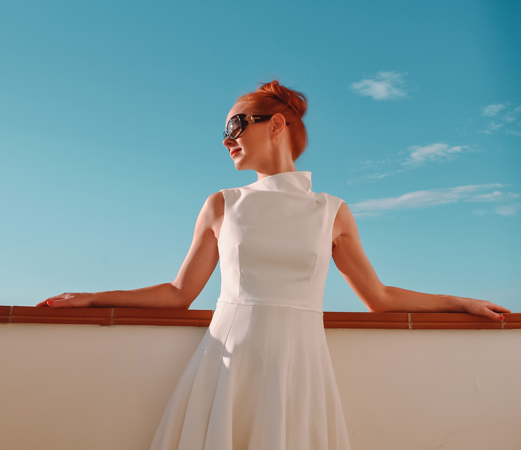 Amber wears a white, high-necked mini dress with a full skirt as she stands on the hotel balcony in the late evening sun