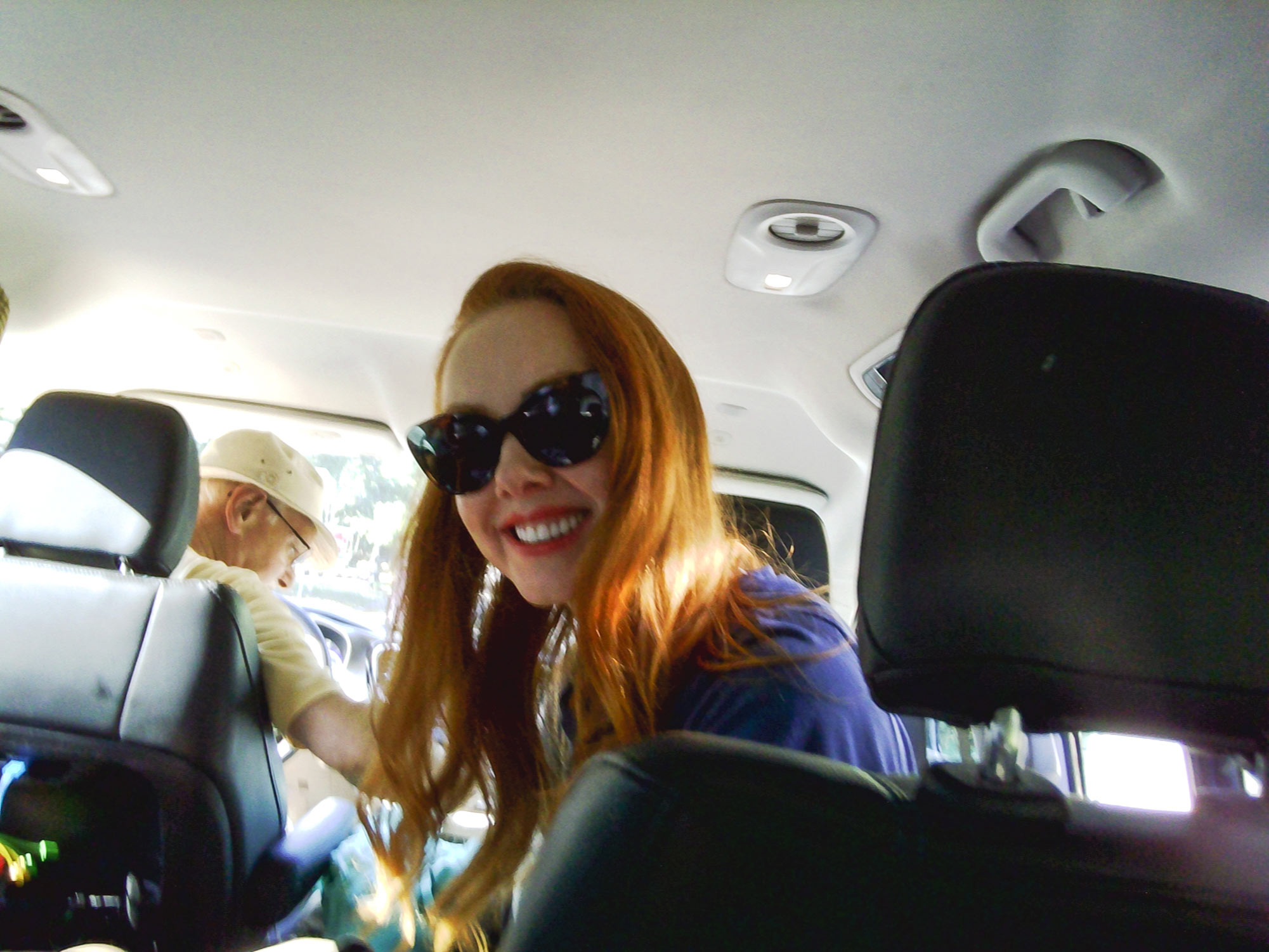 Amber looks round from the passenger sear of a car. She is smiling and wearing dark glasses.